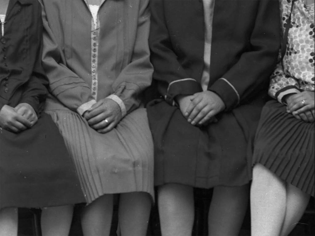 Neljä naista istuu kädet sylissä, päät ja jalat rajattu pois, mustavalkoinen kuva. 