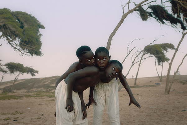 Kolme lasta seisovat toisiinsa nojaten tuulisella savannilla.
