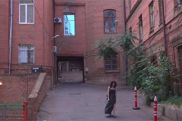 Nainen pitkässä hellemekossa kävelee punatiilisten kerrostalojen sisäpihalta.