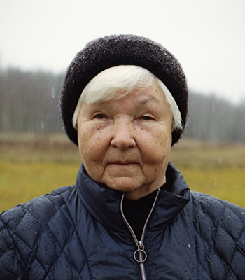 Vanha, hattupäinen nainen katsoo kameraa kohti sateessa vihreä pelto takanaan.