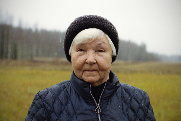 Vanha, hattupäinen nainen katsoo kameraa kohti sateessa vihreä pelto takanaan.