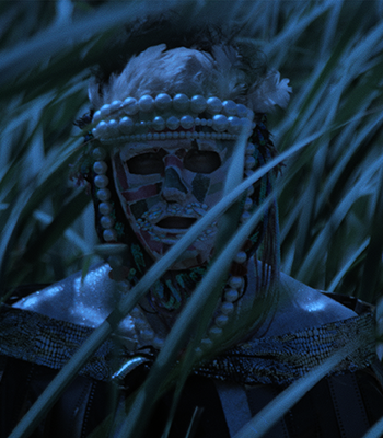 Helmin koristeltu ja värikkääseen maskiin pukeutunut henkilö seisoo pimeässä korkean ruohon seassa.