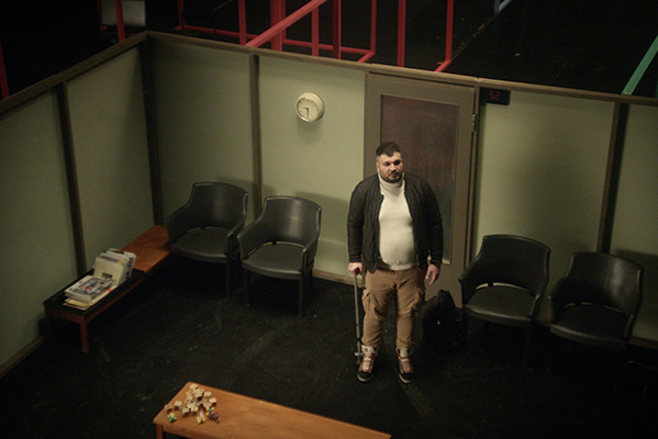 Mies seisoo kävelykepin kanssa lavastemaisessa odotusaulassa.