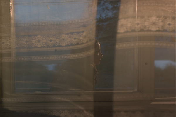 Surumielinen nainen istuu ulkona pitsiverhoilla peitetyn ikkunan takana.