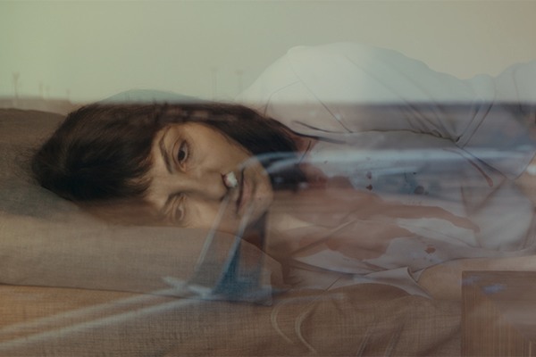 Sängyllä makaa nainen vakavan näköisenä, toisessa sieraimessaan paperituppo verenvuotoa tyrehdyttämään.