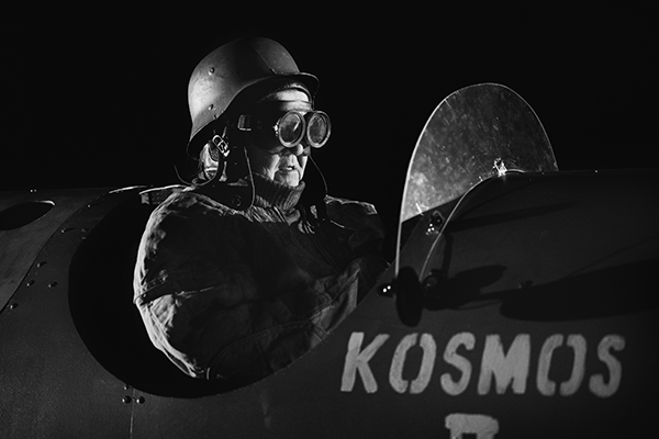 Hahmo vanhanaikaisessa kypärässä ja suojalaseissa lentää vanhaa lentokonetta, jonka kyljessä lukee Kosmos.