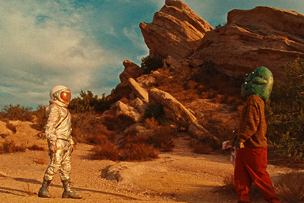 Astronautti ja avaruusoliomainen hahmo seisovat vastakkain aavikkomaisemassa.