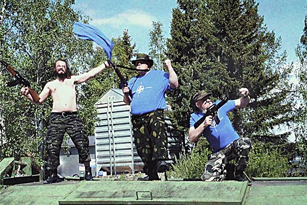 Kolme miestä poseeraavat riehakkaan näköisinä kiväärien kanssa ison kierrätysastian päällä.