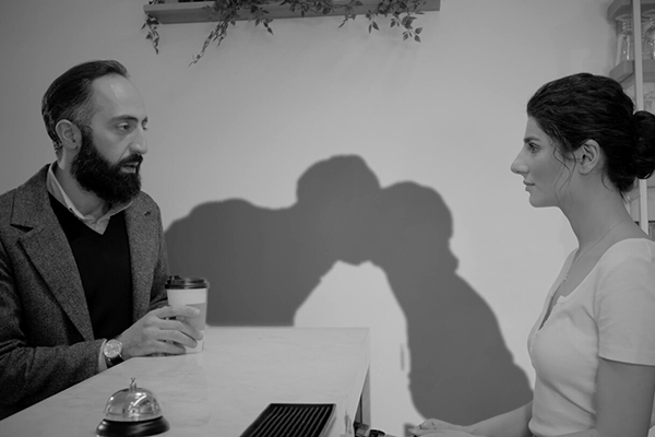 Mies ja nainen istuvat vastakkain pöydän ääressä, heidän varjonsa suutelevat.