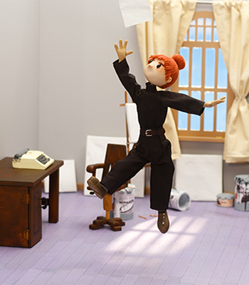 Stop motion -animoitu mustapukuinen hahmo hyppää valkoisen paperiarkin perässä valoisassa ateljeessa.
