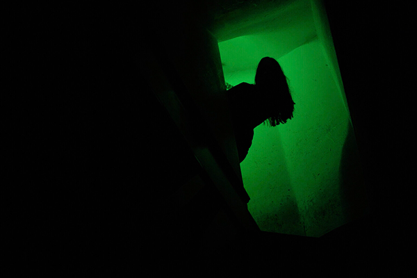 Pimeä huone, josta erottuu vain vihreänä hehkuva oviaukko ja siitä kurkkaava pitkähiuksisen hahmon siluetti.