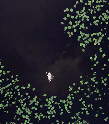 Korkealta otettu kuva tummasta järvestä, jonka keskellä kelluu valkeapukuinen hahmo.