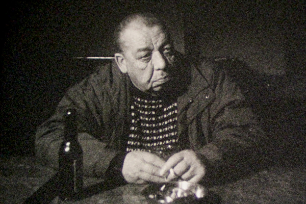 Vanha mies seisoo pöydän äärellä tupakka kädessään ja kaljapullo edessään.