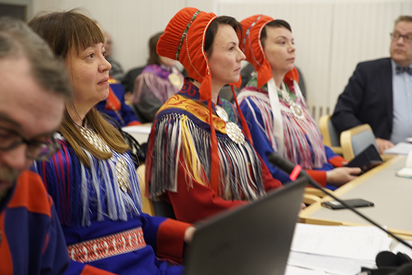 Neljä saamelaisten perinnepukuihin pukeutunutta henkilö istuvat pöydän ääressä.