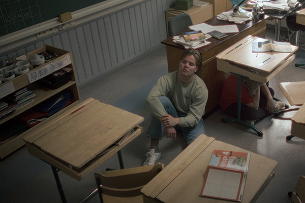 Nuori mies istuu sotkuisen luokkahuoneen lattialla. Hänen vieressään lapsi istuu pulpetin alla.