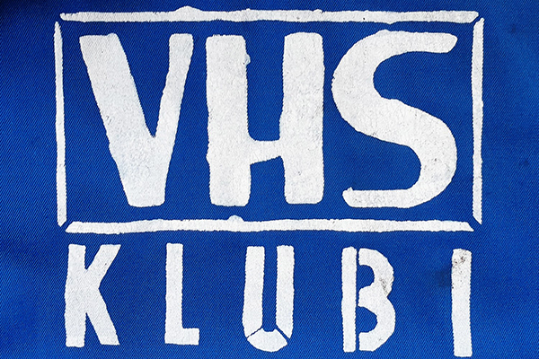 Sininen kangas, johon on painettu valkoisella logo. Logossa lukee isolla "VHS Klubi".