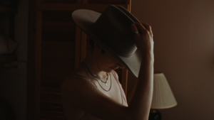 Henkilöllä päässään stetson-hattu, josta hän pitää toisella kädellä kiinni, kasvot painautuneena alaspäin.
